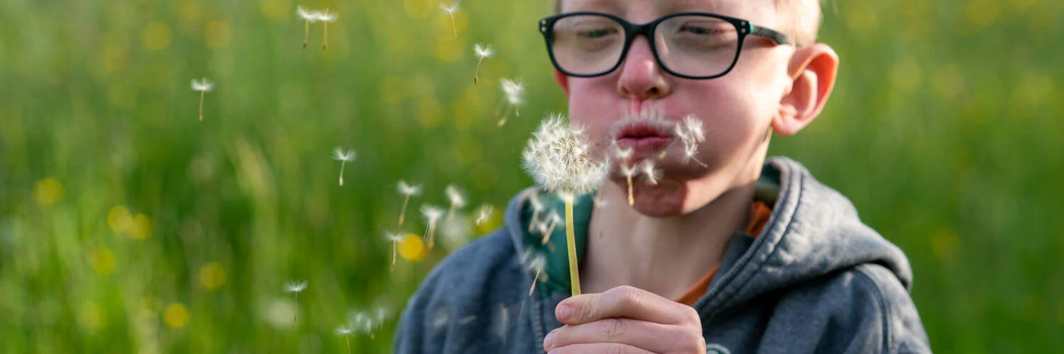 Ein Kind mit Noonan-Syndrom bläst eine Pusteblume {{brizy_dc_image_alt imageSrc=